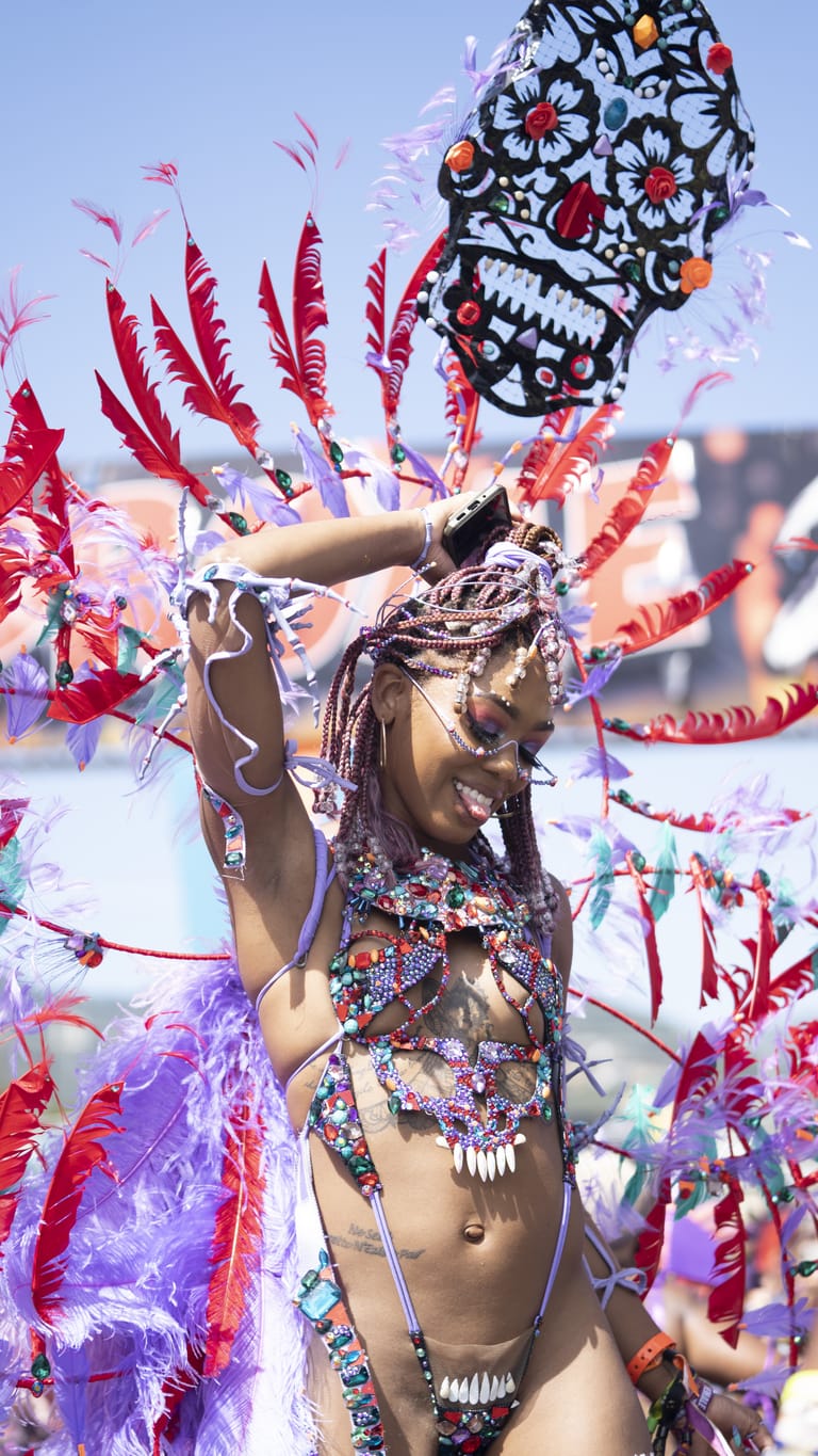 A masquerader in a crimson and violet costume with a tall carved headpiece, phone raised