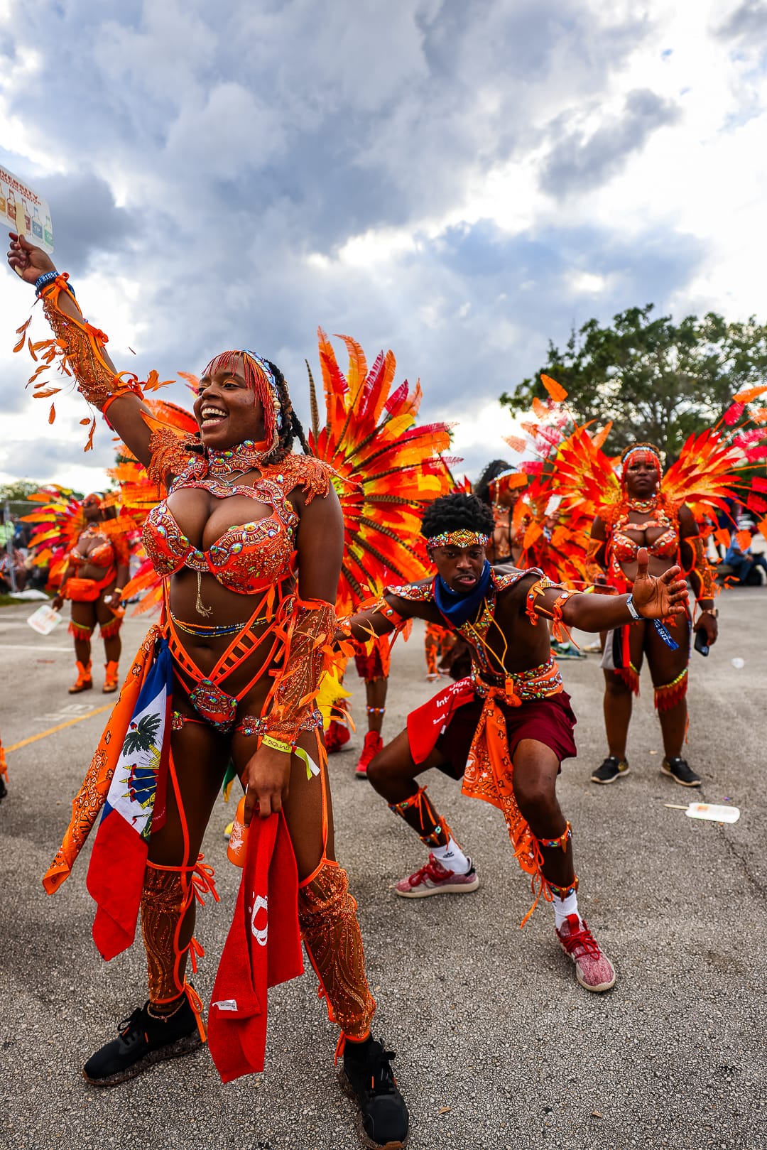 A masquerader in an orange feathered costume holding a flag aloft in daylight on the road
