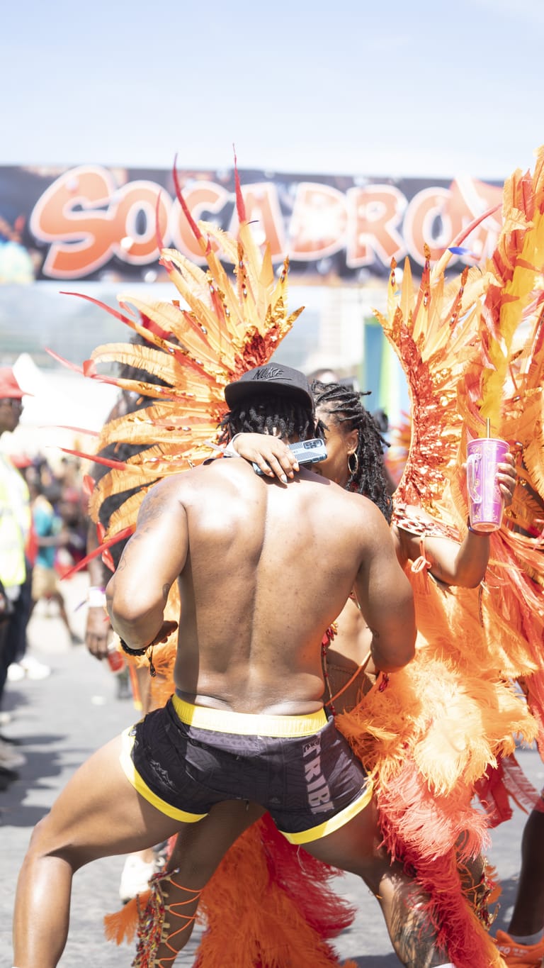 Two masqueraders in orange plumed costumes embrace in front of the Socadrome stage